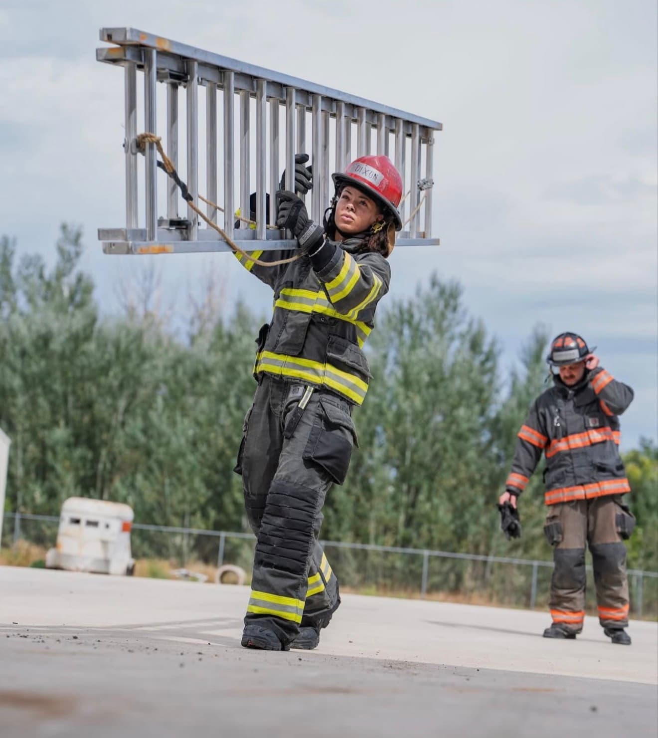 A candidate in full turnout gear raises a ladder overhead during a daylight training evolution