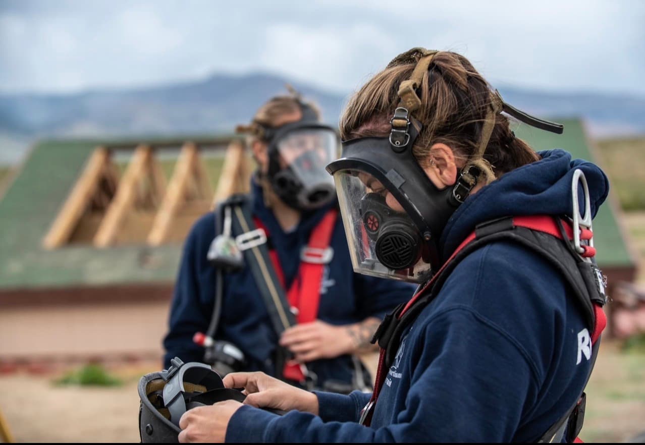 Two women in respirators check equipment between evolutions at a training facility